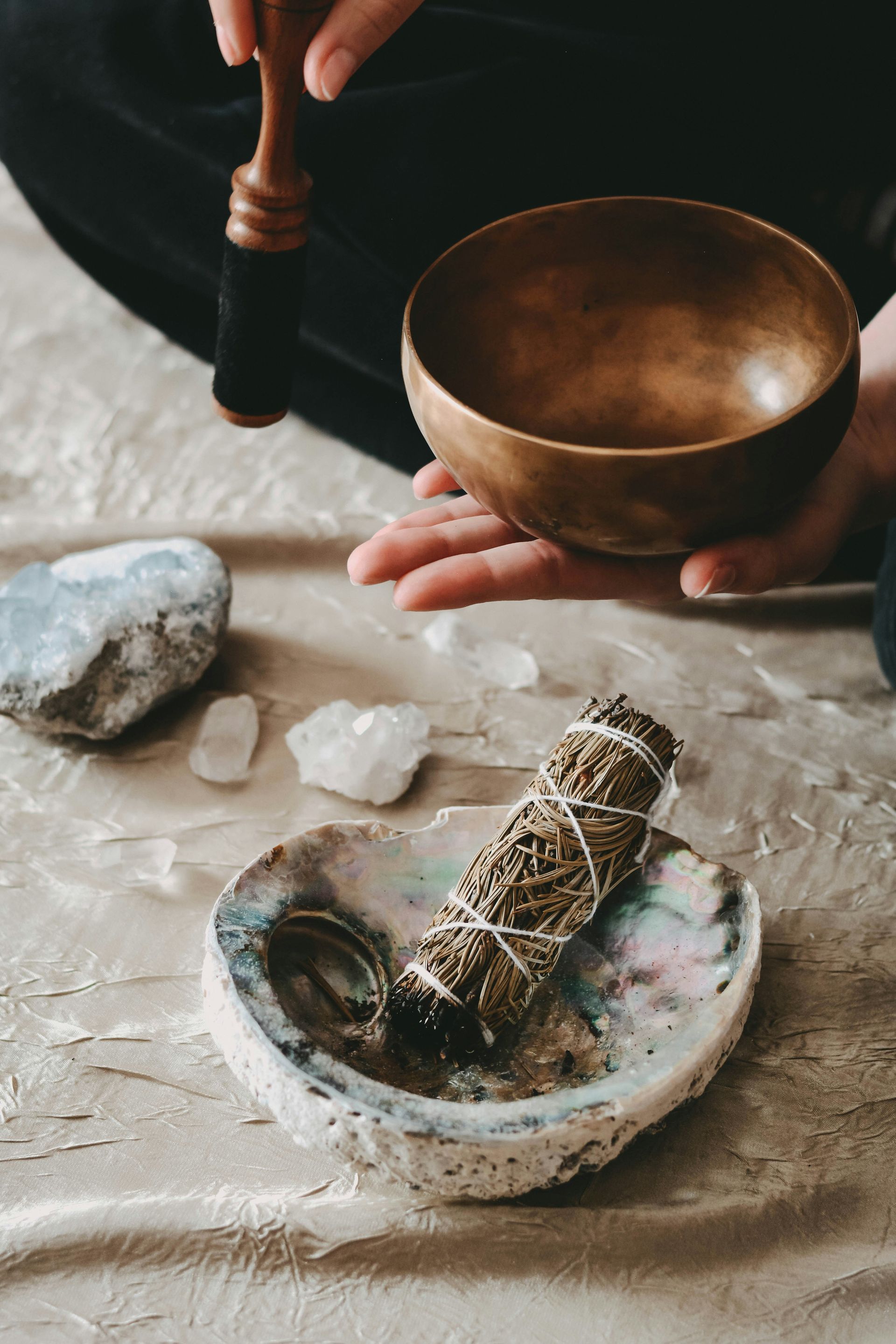 A person holding a bronze singing bowl with a wooden mallet near crystals and sage